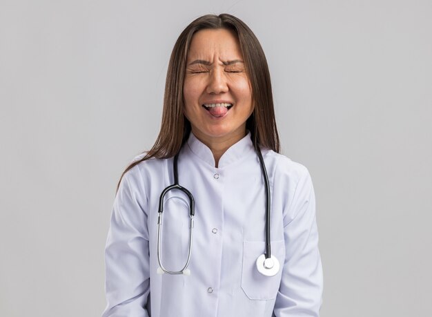 흰 벽에 격리된 닫힌 눈으로 혀를 보여주는 의료 가운과 청진기를 입은 쾌활한 젊은 아시아 여성 의사