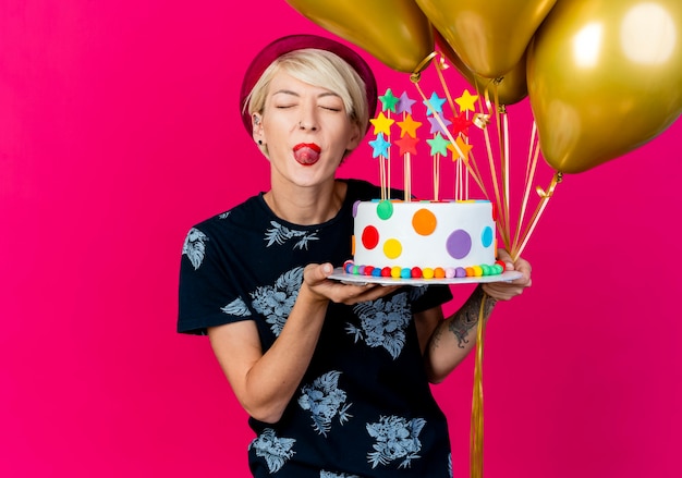 Playful young blonde party girl wearing party hat holding balloons and birthday cake with stars showing tongue with closed eyes isolated on crimson background with copy space
