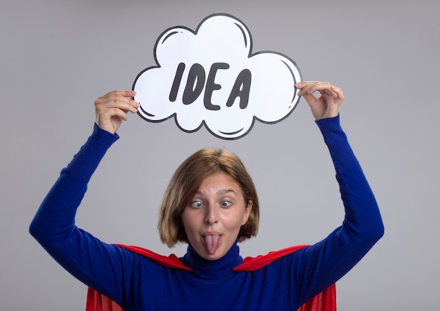 Free photo playful young blonde superhero girl in red cape holding idea bubble above head showing tongue with crossed eyes isolated on white wall