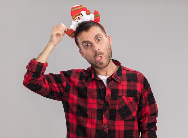 Playful young caucasian man wearing christmas headband grabbing headband looking at camera showing tongue isolated on white background