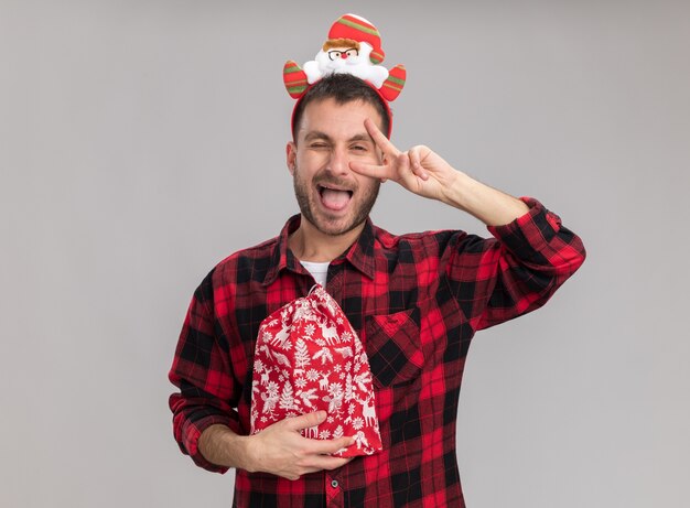 Playful young caucasian man wearing christmas headband holding christmas sack looking at camera showing v-sign symbol near eye winking isolated on white background
