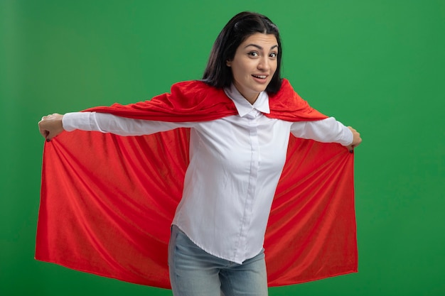 Playful young caucasian superhero girl holding her hero cape and representing flight looking at camera isolated on green background