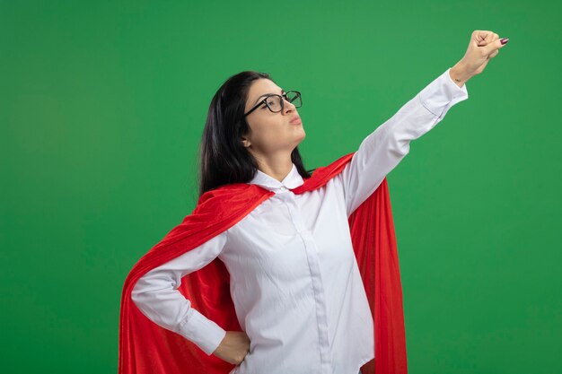 Playful young caucasian superhero girl wearing glasses standing in superman pose in profile view raising her fist up isolated on green background with copy space