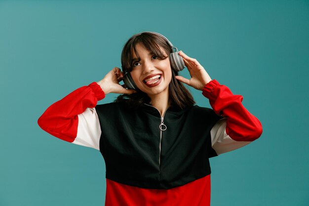 Playful young caucasian woman wearing and grabbing headphones looking up showing tongue isolated on blue background