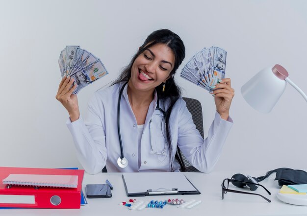 Playful young female doctor wearing medical robe and stethoscope sitting at desk with medical tools holding money showing tongue with closed eyes isolated