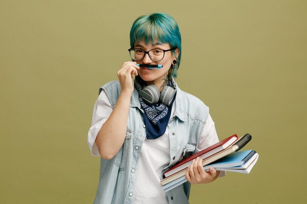 Игривая молодая студентка в очках, бандане и наушниках на шее, держа блокноты, глядя на камеру, делая усы с ручкой, изолированной на оливково-зеленом фоне