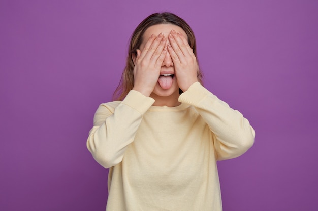 Giocosa giovane ragazza che copre gli occhi con le mani che mostrano la lingua