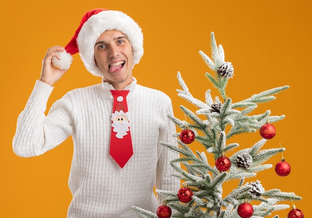 Игривый молодой красивый парень в новогодней шапке и галстуке санта-клауса стоит возле украшенной елки, хватая шляпу, глядя в камеру, показывая язык, изолированный на оранжевом фоне