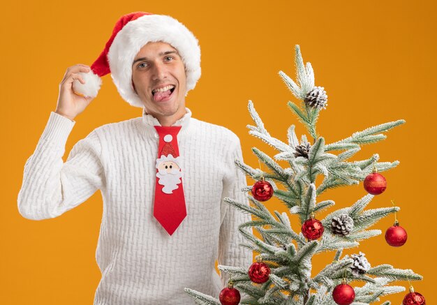 クリスマスの帽子とサンタクロースのネクタイを身に着けている遊び心のある若いハンサムな男は、オレンジ色の背景に分離された舌を示すカメラを見て装飾されたクリスマスツリーをつかむ帽子の近くに立っています