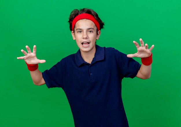 Playful young handsome sporty boy wearing headband and wristbands with dental braces looking at camera doing tiger roar and paws gesture isolated on green background