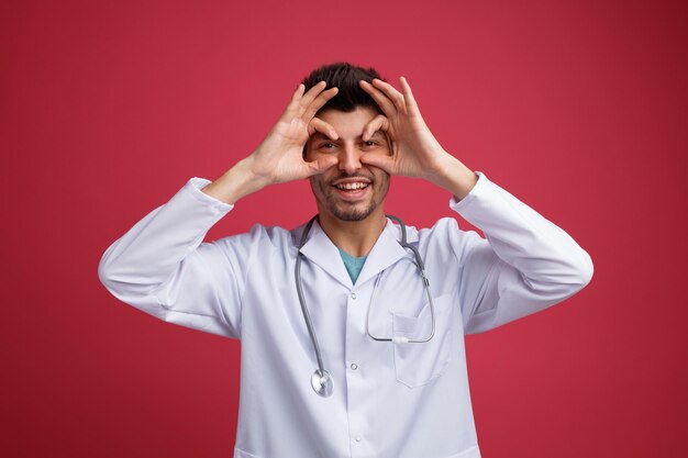 Playful young male doctor wearing medical uniform and stethoscope around his neck looking at camera showing binoculars gesture isolated on red background