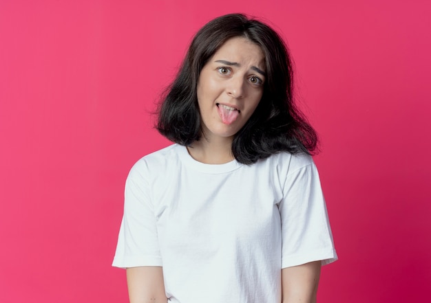 Playful young pretty caucasian girl looking at camera and showing tongue isolated on crimson background with copy space