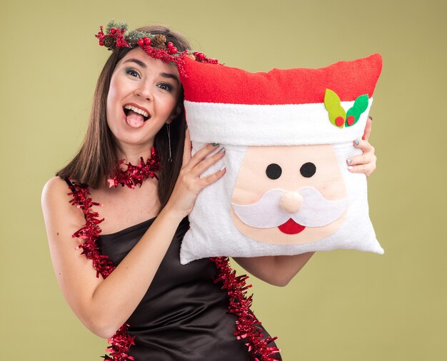 Playful young pretty caucasian girl wearing christmas head wreath and tinsel garland around neck holding santa claus pillow looking at camera showing tongue isolated on olive green background