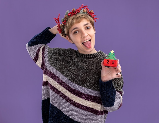 Free photo playful young pretty girl wearing christmas head wreath holding christmas tree toy with date looking at camera keeping hand behind head showing tongue isolated on purple background