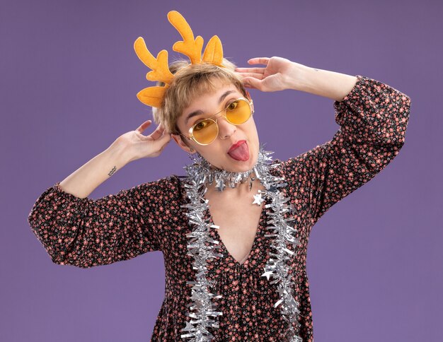 Playful young pretty girl wearing reindeer antlers headband and tinsel garland around neck with glasses looking at camera grabbing headband showing tongue isolated on purple background