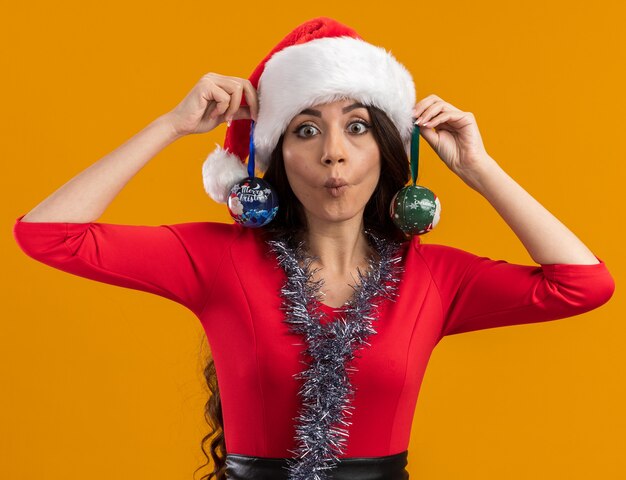 Playful young pretty girl wearing santa hat and tinsel garland around neck hanging christmas baubles on ears looking at camera making fish face isolated on orange background