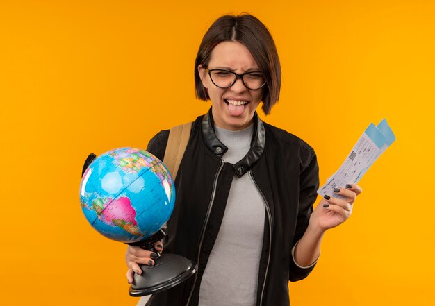 Playful young student girl wearing glasses and back bag holding airplane tickets and globe showing tongue isolated on orange background