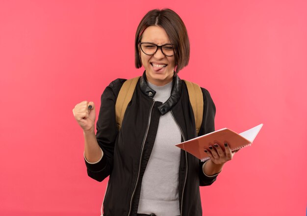 Игривая молодая студентка в очках и задней сумке держит блокнот, показывая язык со сжатым кулаком и закрытыми глазами, изолированными на розовом фоне с копией пространства