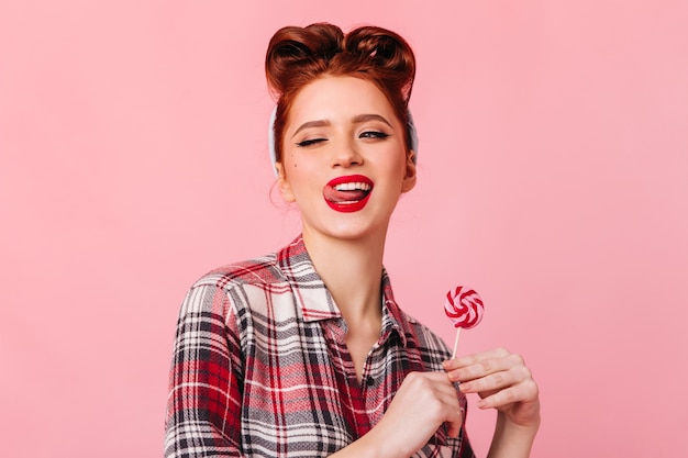Игривая молодая женщина в клетчатой рубашке позирует с конфетой. Потрясающая девушка кинозвезды, стоящая на розовом пространстве с леденцом на палочке.