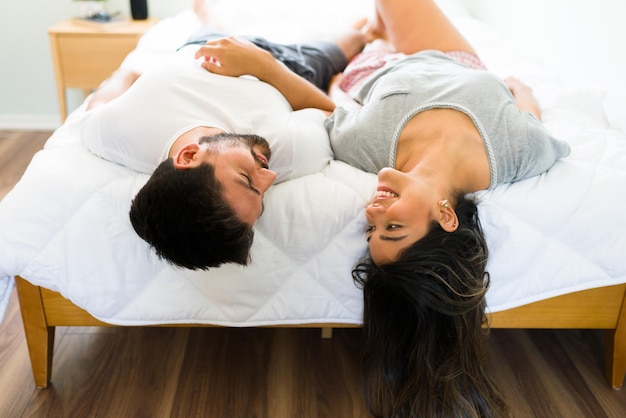 Free photo playful young woman and man in love lying upside down in their comfy bed and talking about happy positive emotions while having fun