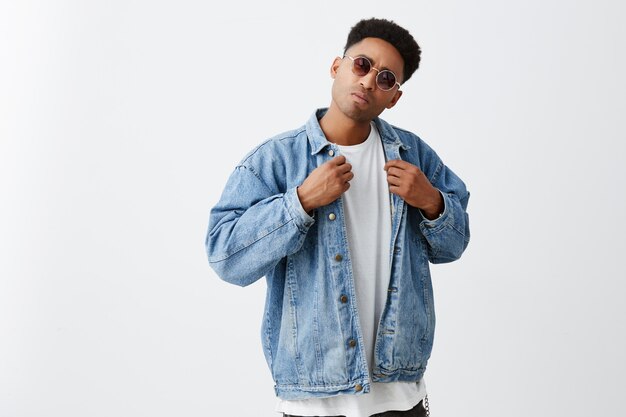 Playing cool. Close up of young good-looking black american man with afro hairstyle in casual white t-shirt, denim jacket and sun glasses looking in camera with mean expression.