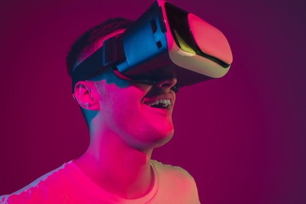 Playing with VR, shoting, driving. Caucasian man's portrait isolated on pink-purple  wall in neon light. Male model with devices. Concept of human emotions, facial expression,