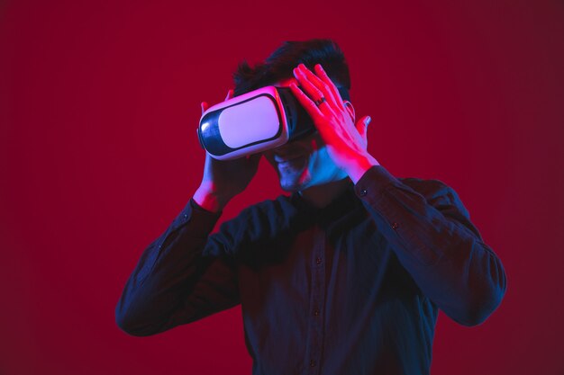 VR 헤드셋을 착용했습니다. 네온 불빛에 붉은 벽에 고립 된 백인 젊은 남자의 초상화. 아름다운 모델. 인간의 감정, 표정, 청소년, 장치의 개념.