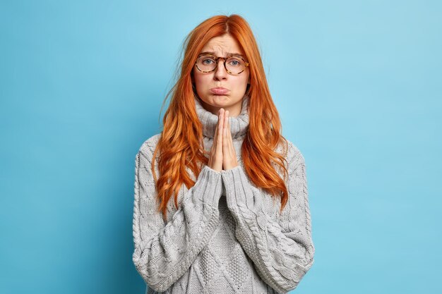 Молящая серьезная рыжая женщина с расстроенным выражением, сжимающая ладони в молитве, недовольна, одетая в повседневный серый свитер.