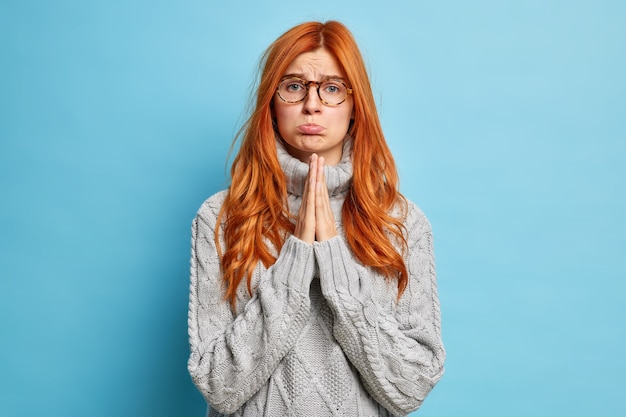 Pleading serious ginger woman upset expression keeps palms pressed together in prayer has dissatisfied expression dressed in casual grey sweater.