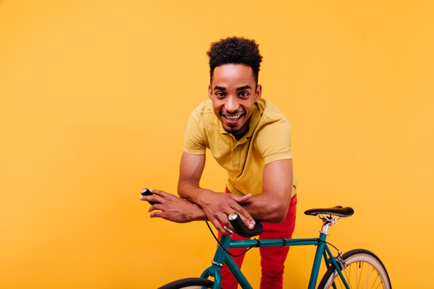 Pleasant african man with curly hair standing beside bicycle. Indoor shot of ecstatic black guy posing