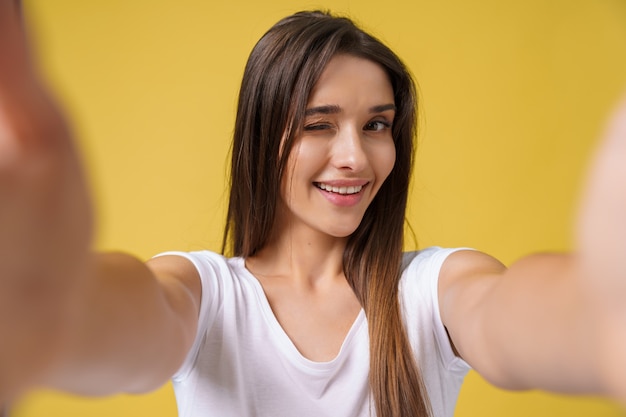 Free photo pleasant attractive girl making selfie in studio and laughing. good-looking young woman with brown hair taking picture of herself on bright yellow background.