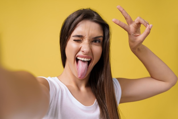 Piacevole ragazza attraente che fa selfie in studio e ride bella giovane donna con i capelli castani che scatta foto di se stessa su sfondo giallo brillante