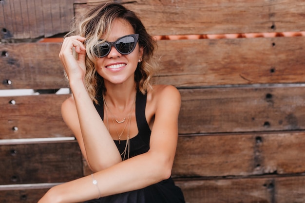 Pleasant blonde woman posing in dark sunglasses on wooden wall. attractive caucasian female model in black clothes smiling