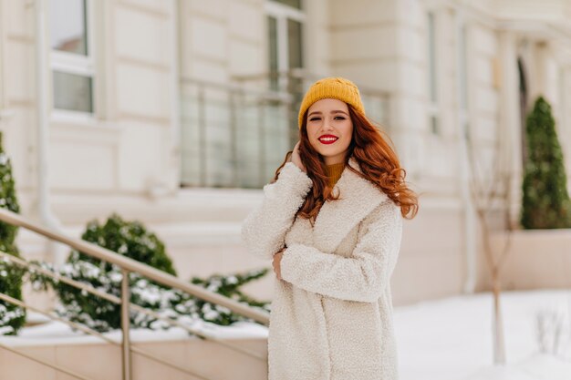 Приятная кавказская женщина в белом халате наслаждается прогулкой на выходных. Открытый портрет стильной имбирной девушки в зимнем наряде.