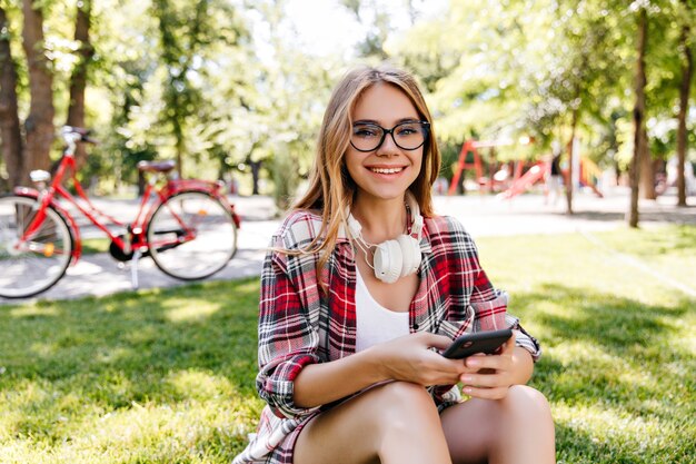 芝生の上に座って電話を使用して快適なヨーロッパの女性モデル。公園でスマートフォンでポーズをとる白人のかっこいい女の子。
