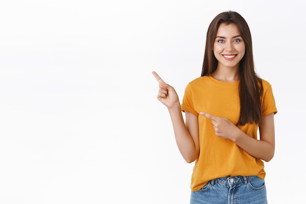 左上隅を指している黄色のTシャツの快適なフレンドリーな見た目のかなり白人女性は、顧客が自信を持って立っている白い背景に立っている製品やイベントのプロモーションをお勧めする方法を見つけるのに役立ちます