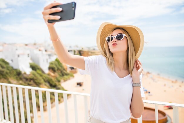 Pleasant girl in sunglasses and hat make kiss on camera while making selfie on sea