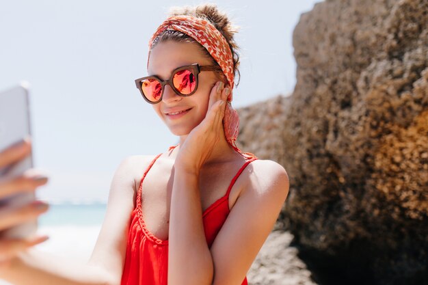 Pleasant girl in sunglasses touching her cheek while making selfie in the beach. Outdoor photo of inspired tanned woman chilling at beach and taking pictures of herself.