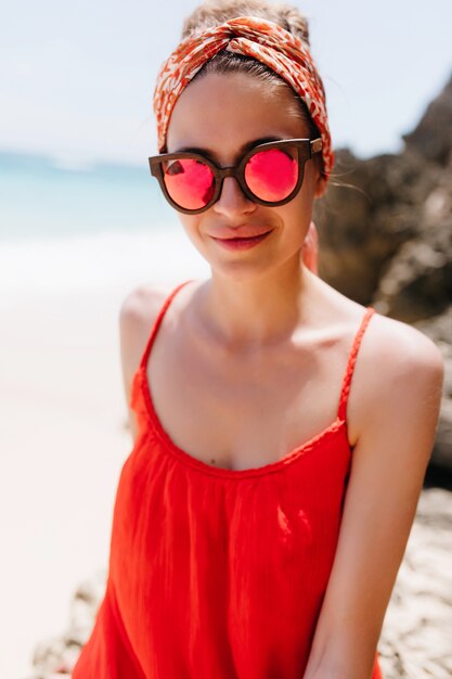 ビーチでポーズをとる流行の服装の楽しい女の子。海岸を歩いているピンクのサングラスで魅力的な日焼けした女性の屋外の写真。