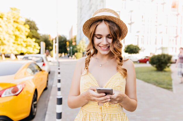 通りに立ってメッセージをテキストメッセージで送る楽しい魅力的な女の子。スマートフォンでポーズをとってレトロな帽子の愛らしい女性の屋外の肖像画。