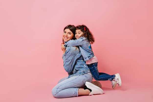 Pleasant kid embracing mother on pink background. Studio shot of blissful mom and little daughter in jeans.