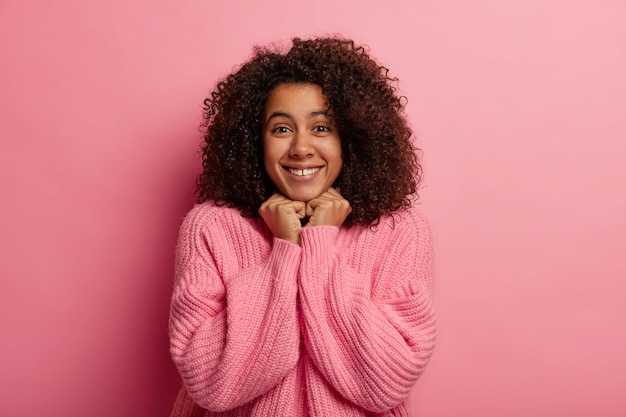 見栄えの良いアフロの女性は優しく微笑んで、両手をあごの下に置き、健康な肌を持ち、冬のセーターを着て、前向きなニュースを受け取り、ピンクの壁に隔離されています。