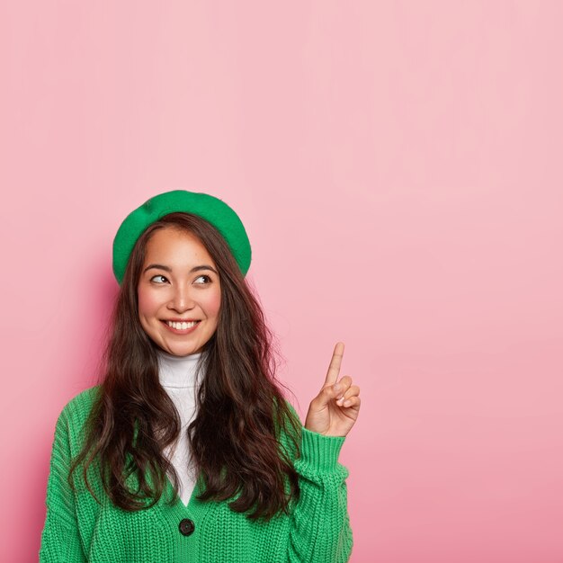 유쾌한 아시아 여성은 녹색 베레모와 니트 점퍼를 입고 앞쪽 손가락을 가리키며 쾌활한 표정을 지닙니다.