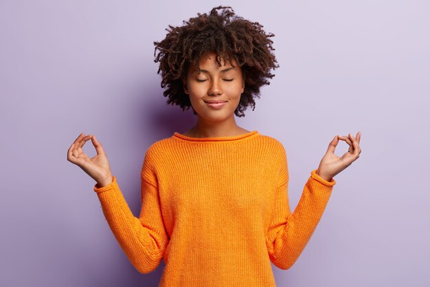 Pleasant looking calm woman meditates indoor, holds hands in mudra gesture, has charming smile, closed eyes, wears orange clothes, models over purple wall. Hand gesture. Meditation concept