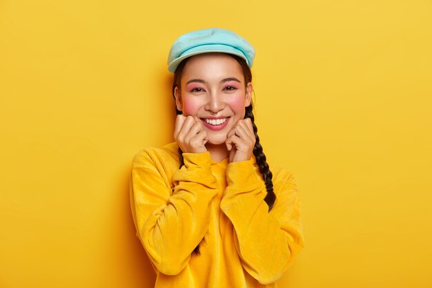 Pleasant looking cheerful Asian girl keeps both hands under chin, has pinup makeup, wears blue stylish cap, velvet yellow hoodie