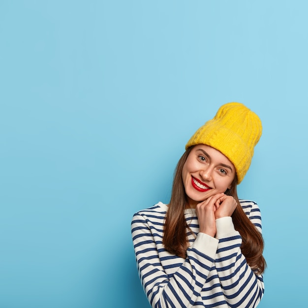 Бесплатное фото Приятная на вид жизнерадостная женщина наклоняет голову, ласково улыбается, держит руки вместе у подбородка, носит желтую шляпу и полосатый матросский джемпер.