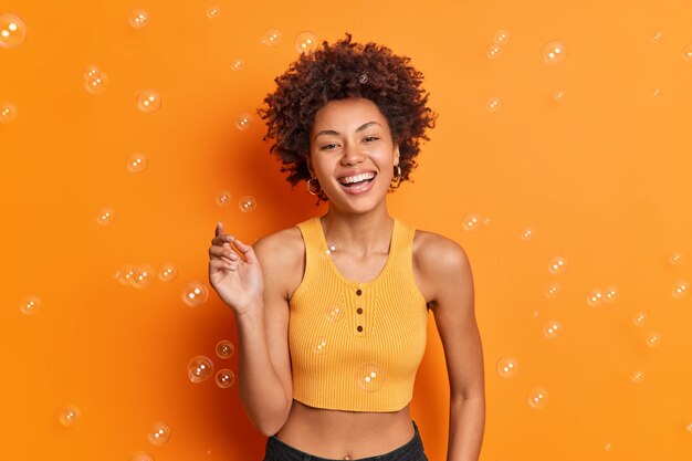 Pleasant looking cheerful young Afro American teenage girl keeps hand raised smiles broadly has carefree expression wears casual clothes poses against orange wall with flying soap bubbles