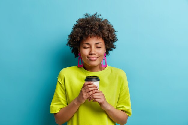 Pleasant looking curly young woman closes eyes enjoys takeaway coffee feels pleasure has spare time wears casual t shirt and pink earrings isolated over blue wall