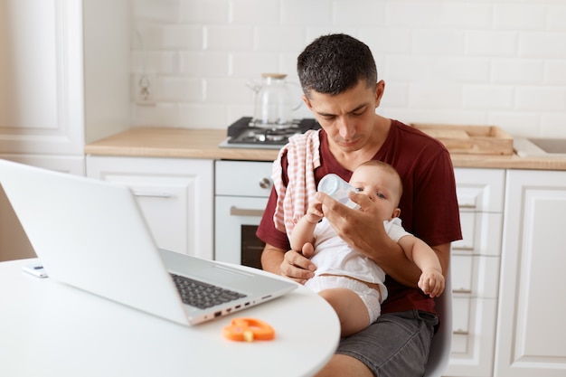 Pleasant looking dark haired handsome man wearing casual t shirt with towel on his shoulder, sitting at table with laptop, holding baby girl in hands, giving baby water to drink.