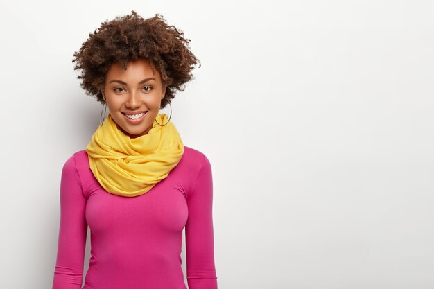 Pleasant looking fashionable woman with Afro hairstyle, wears pink sweatshirt and yellow scarf, smiles happily